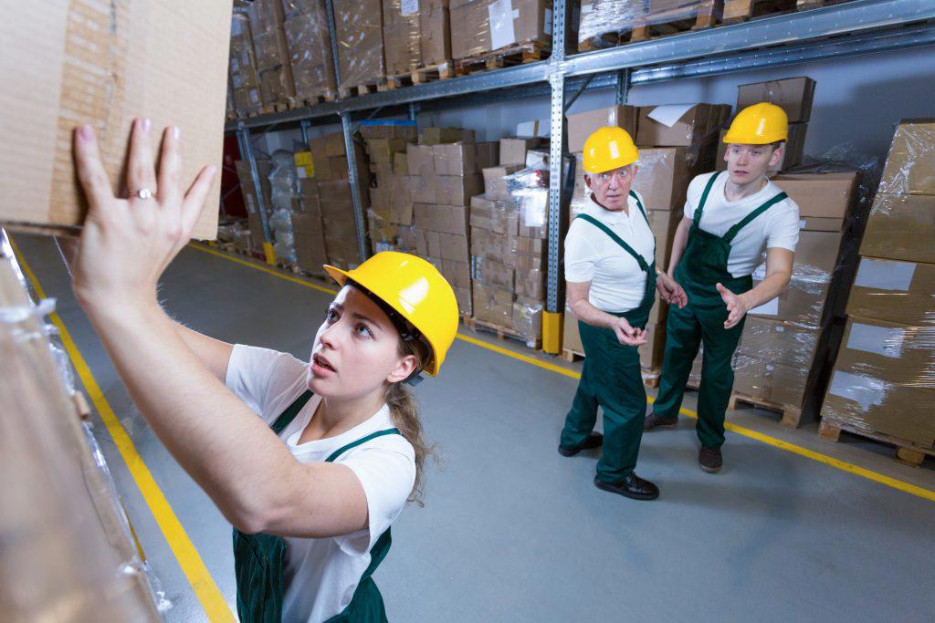 woman worker struggling to put heavy box away up high with coworkers worried watching in the background
